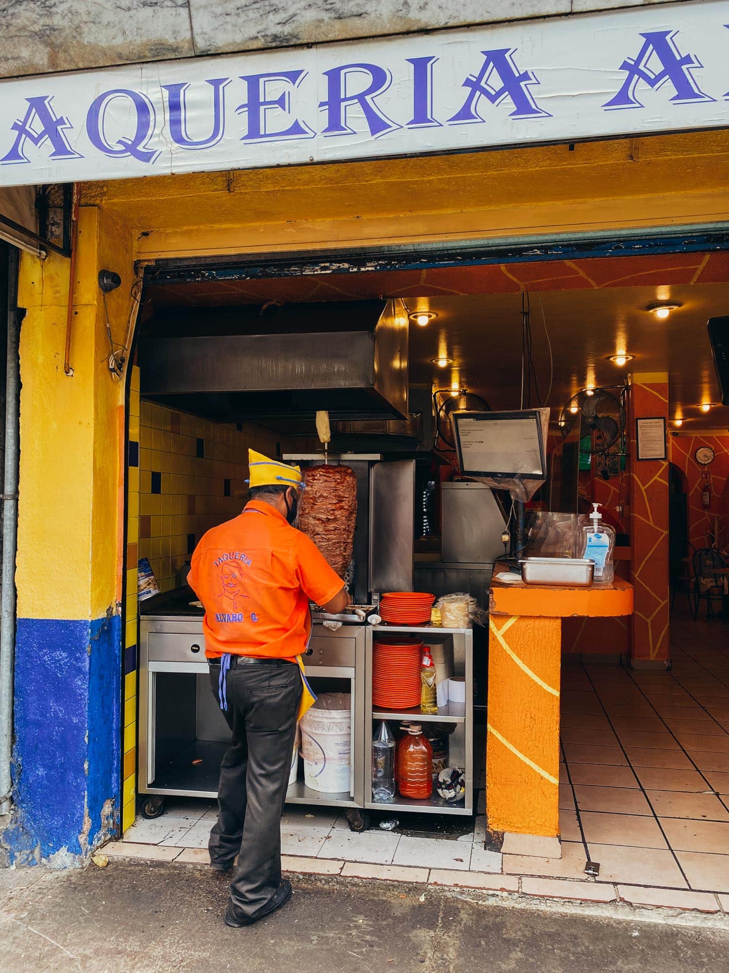 A taqueria worker in an orange shirt slices meat from a vertical rotisserie for tacos al pastor inside a colorful eatery with the sign 'Taqueria Alvaro Obregon' overhead.