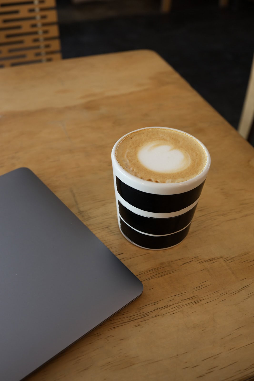 A latte served in a black and white striped cup, placed on a wooden table next to a sleek, silver laptop. The setup gives a cozy, modern vibe perfect for working in a café.