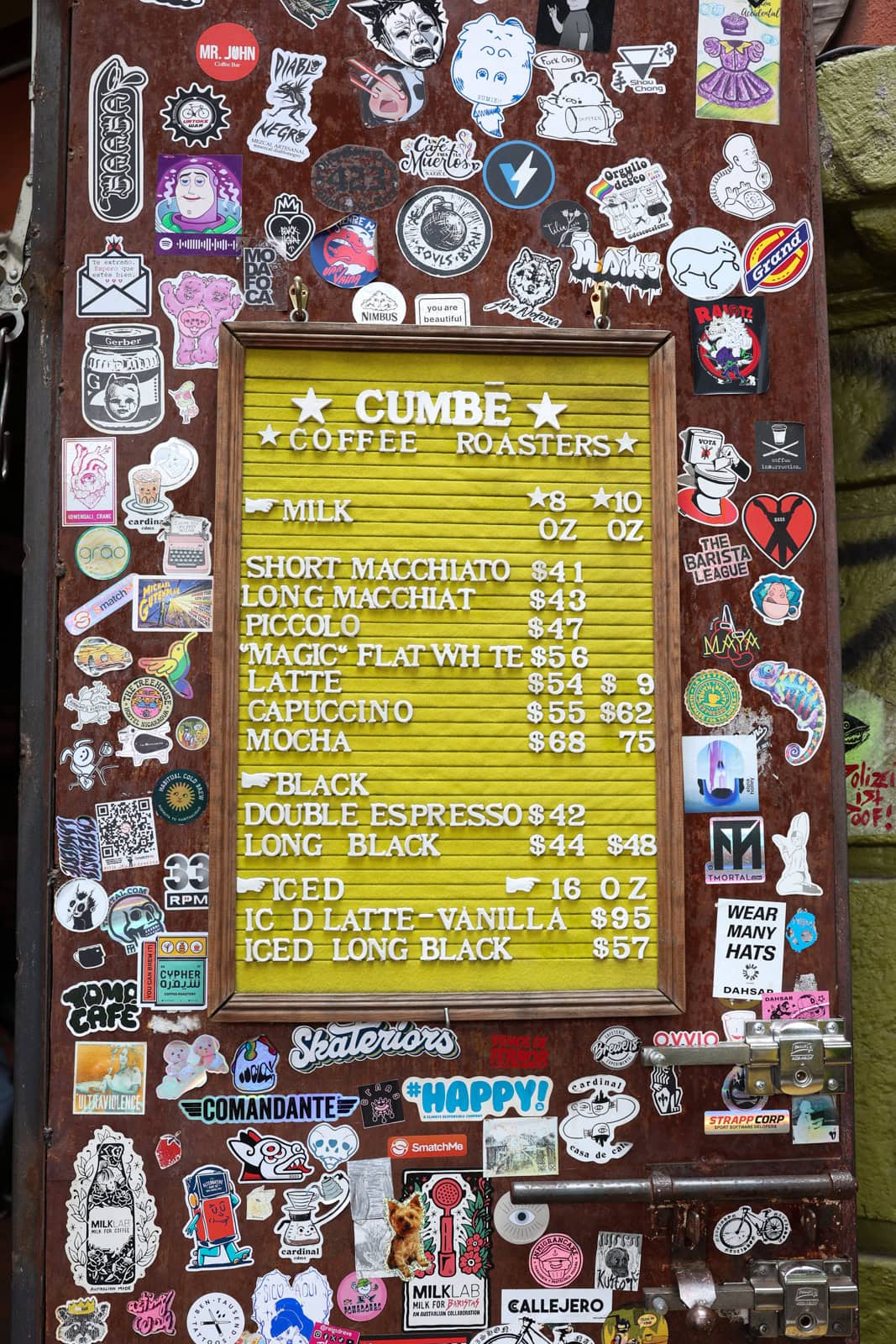 A menu board for 'CUMBÉ Coffee Roasters' displaying a variety of coffee options, surrounded by numerous colorful stickers on a rustic wooden door.