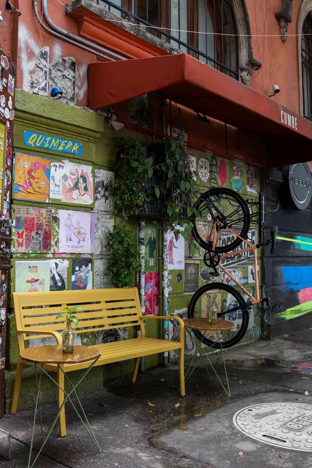 A small outdoor seating area with a yellow bench and matching yellow tables and chairs. The wall behind is covered with colorful posters and stickers, and a bicycle is mounted on the wall as decoration.