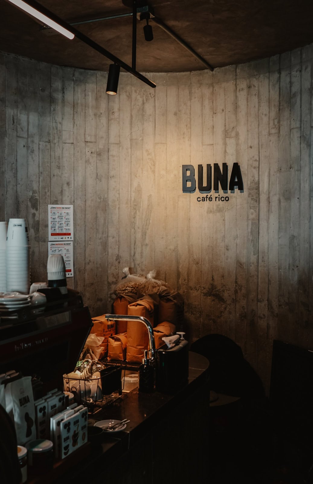 Interior of a coffee shop with a minimalist, industrial design featuring a concrete wall with the words 'Buna café rico' in bold black letters. The counter is cluttered with coffee equipment, bags of coffee beans, and other supplies, giving the space a cozy, artisanal atmosphere.