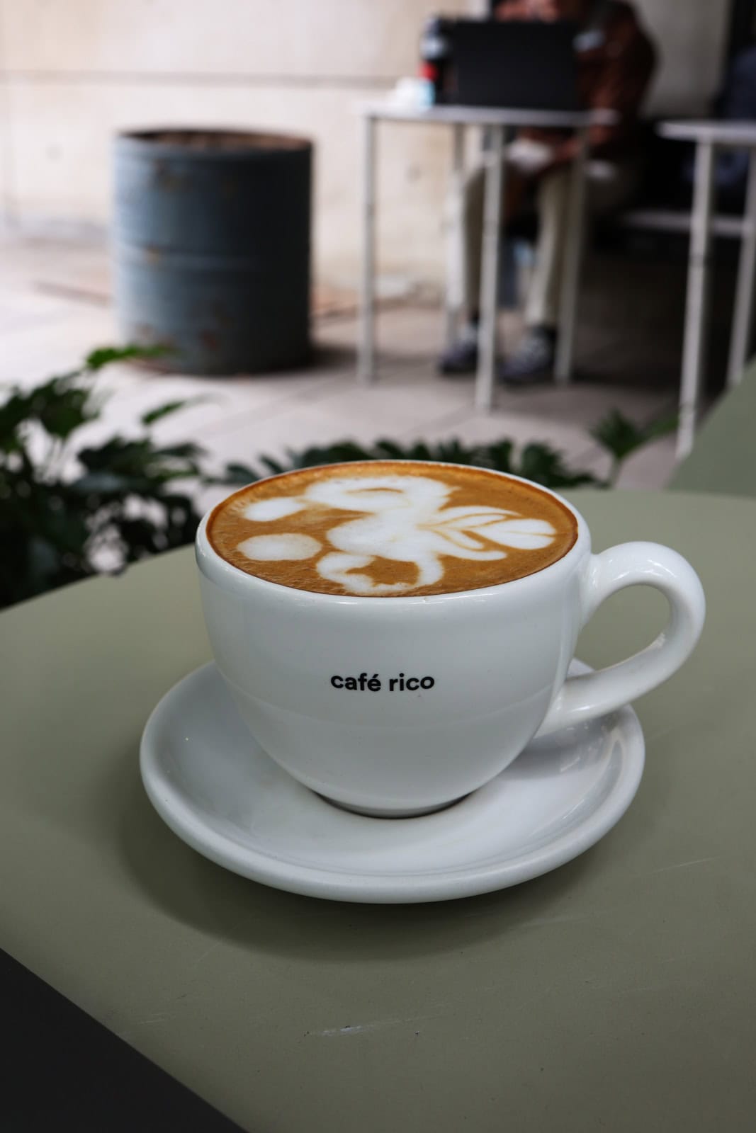 A white coffee cup with the words 'café rico' printed on it, sitting on a saucer. The cup contains a latte with intricate foam art on top, placed on a light green table in an outdoor setting.
