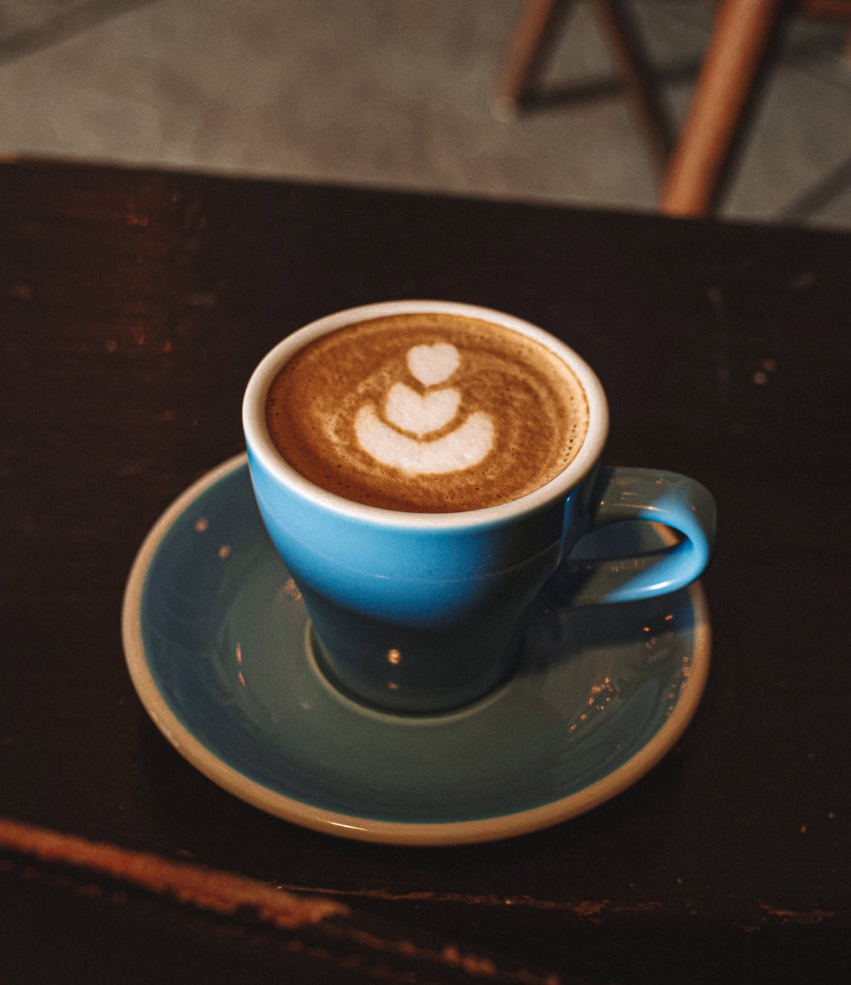 A cappuccino served in a blue cup with matching saucer, placed on a dark wooden table. The foam on the cappuccino is carefully crafted into a simple yet elegant design.