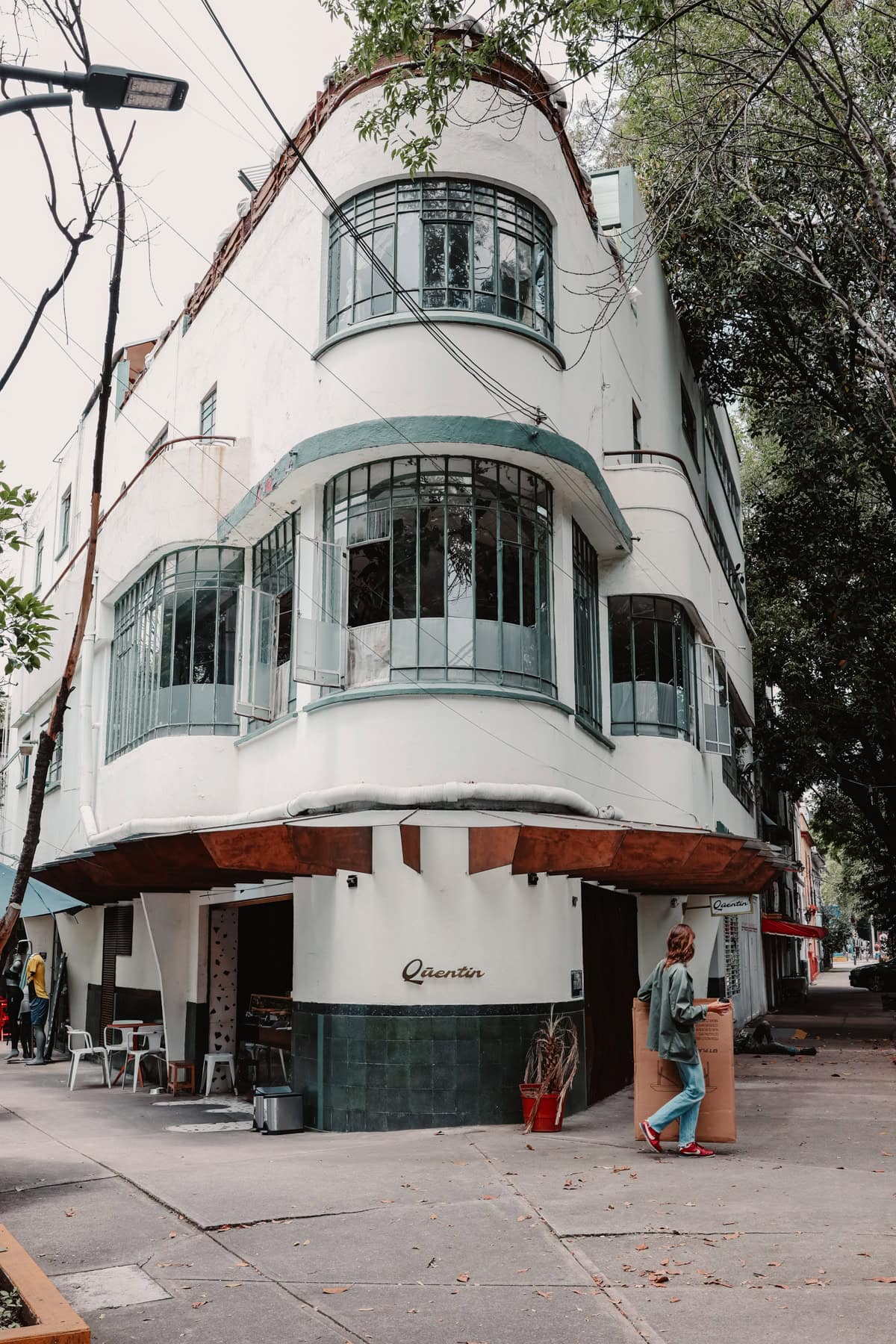 A street view of a corner building with an art deco architectural style, featuring large, curved windows and a small cafe named Quentin on the ground floor. A person is walking past the cafe holding a large cardboard box.