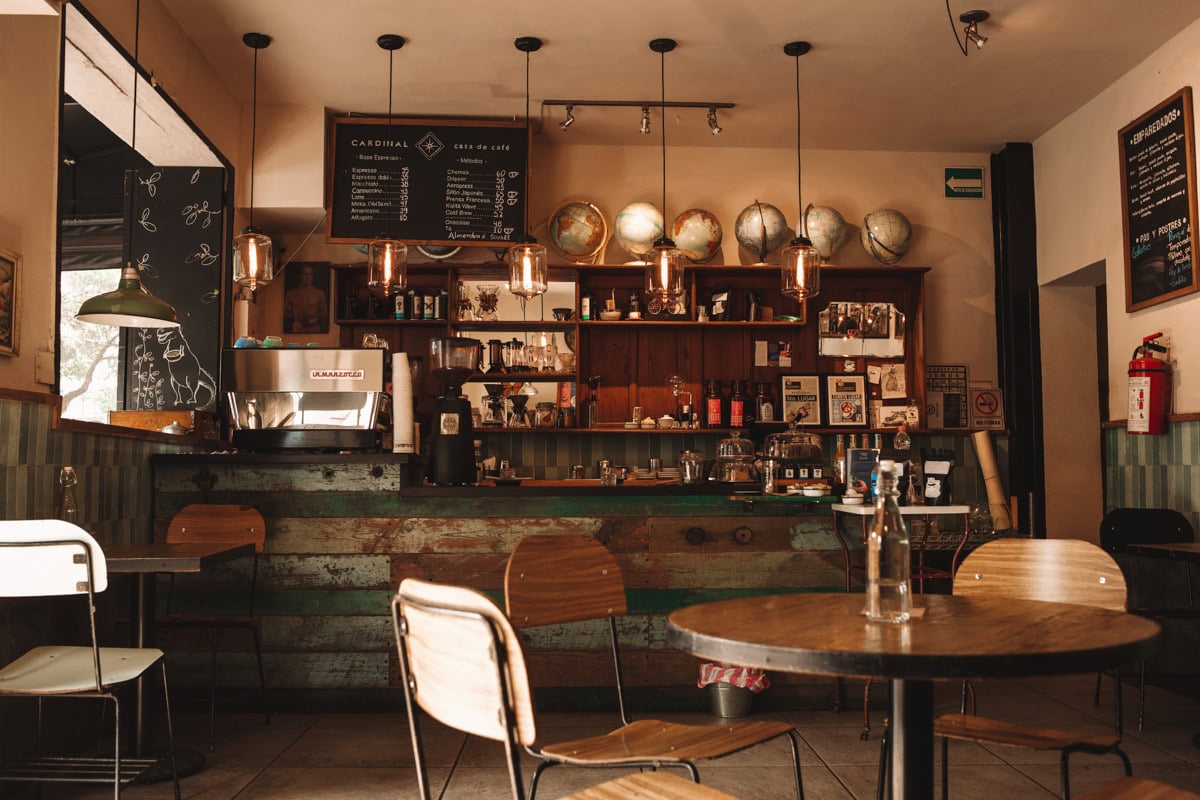 The interior of Cardinal Coffee Shop, featuring a rustic and eclectic design. The counter is made from reclaimed wood, with shelves behind it holding coffee equipment and a collection of vintage globes. The space is warmly lit by pendant lights, with a menu board overhead and a mix of wooden chairs and tables in the seating area.