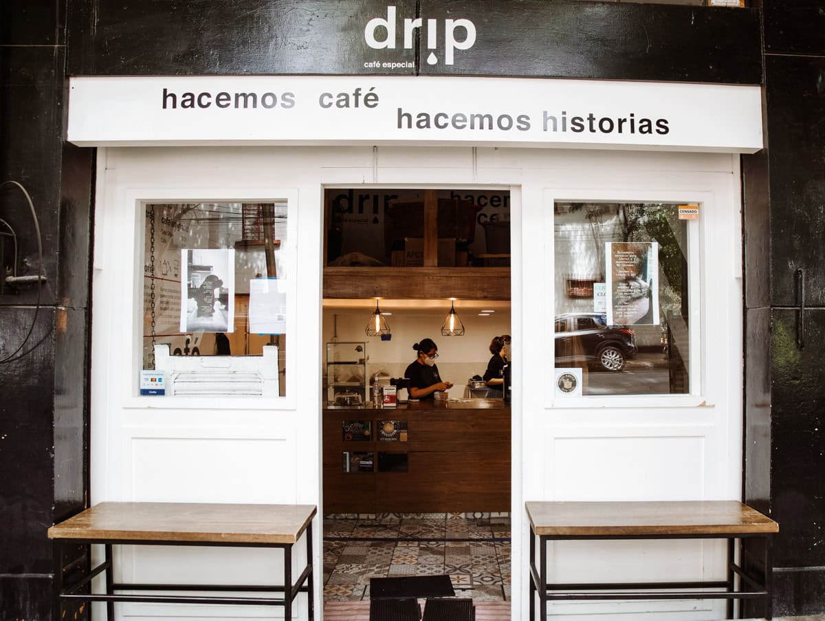 The exterior of Drip Coffee Shop with a clean, minimalist design. The storefront features white double doors with large windows, through which you can see staff working behind the counter. The sign above reads 'drip café especial' with the words 'hacemos café, hacemos historias' written across the facade.