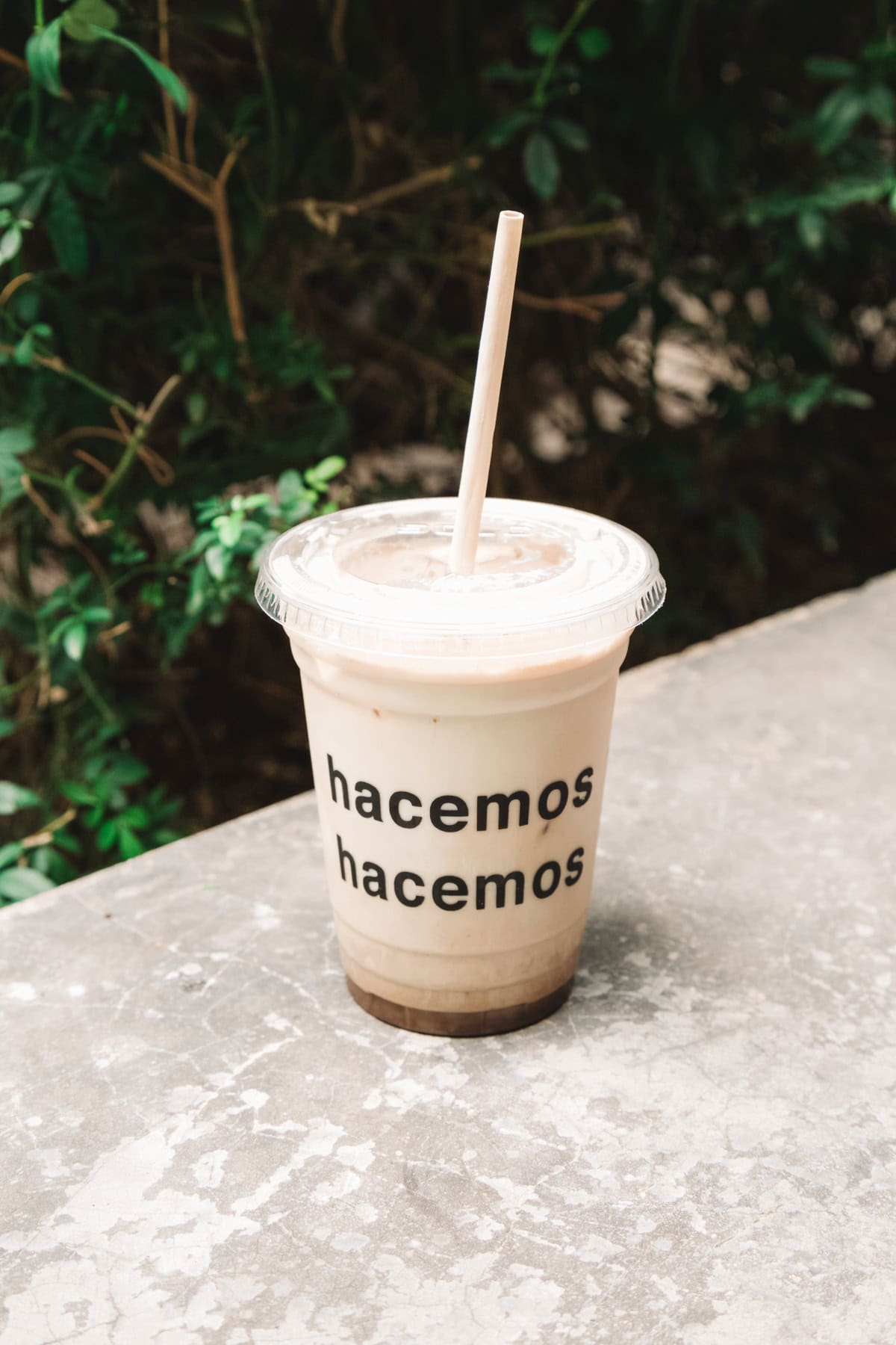 A plastic cup of iced coffee from Drip Coffee Shop with the words 'hacemos hacemos' printed repeatedly in black on the cup. The cup is placed on a textured stone surface with green foliage in the background.