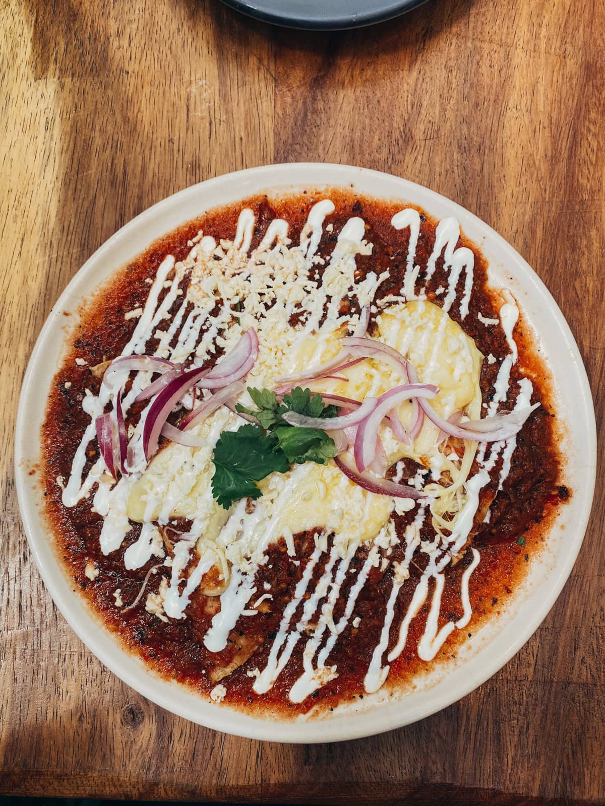A serving of enchiladas with green sauce, topped with crumbled queso fresco and cilantro, on a plastic green plate, surrounded by condiments and drinks on a casual street food table setup.