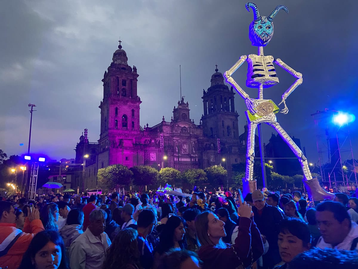 A giant skeleton sculpture with a blue demon mask, standing in front of a lit-up cathedral in Mexico City during a Day of the Dead celebration. A crowd of people gathers below, many taking photos and enjoying the event.