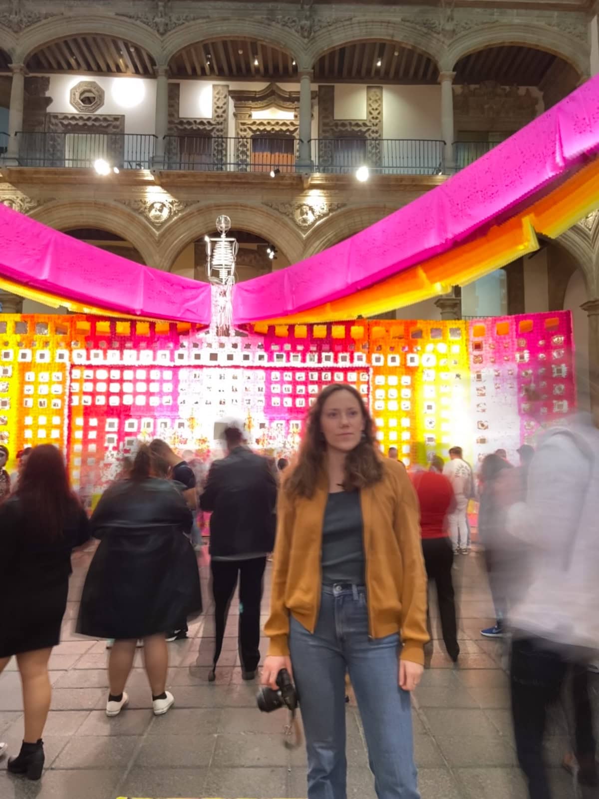 A colorful Day of the Dead altar inside a historic building, decorated with bright pink, orange, and yellow papel picado banners. A woman in casual attire holding a camera stands in the foreground as blurred figures pass by.