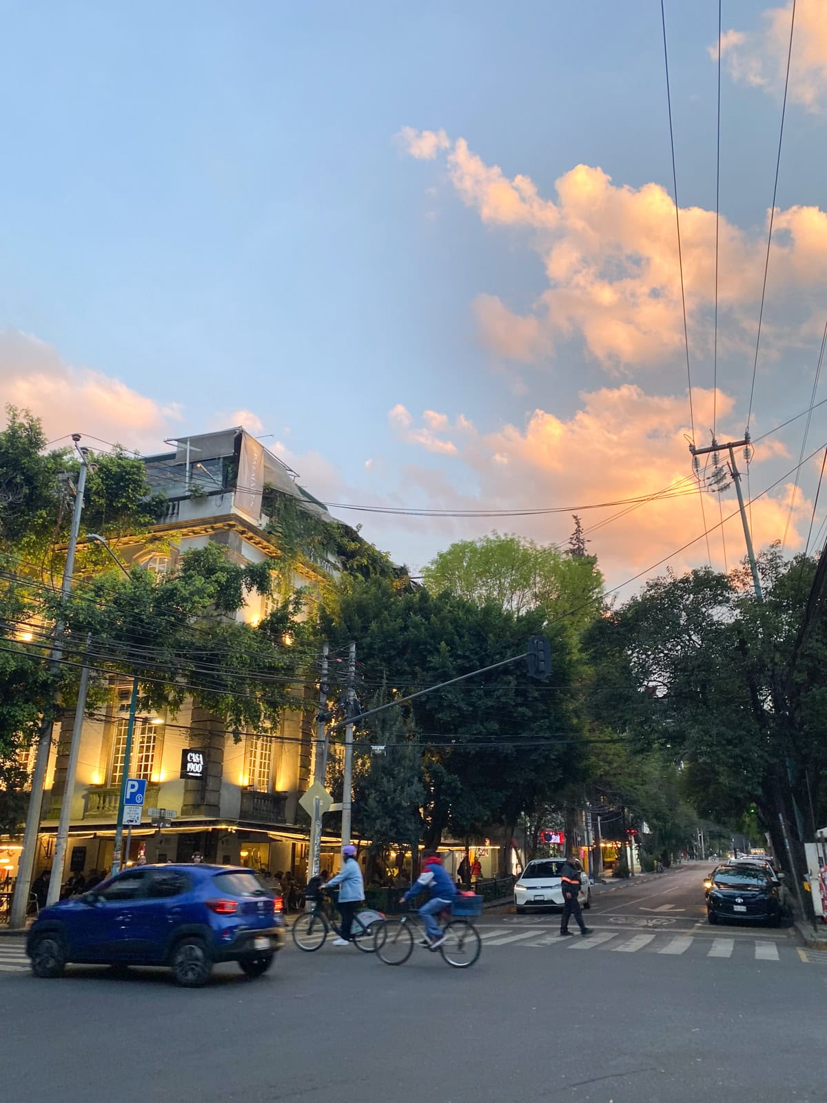 A lively city street at sunset with golden light reflecting off a building covered in greenery. Cyclists and pedestrians cross the intersection, while cars and power lines add to the urban scene under a sky filled with glowing clouds.