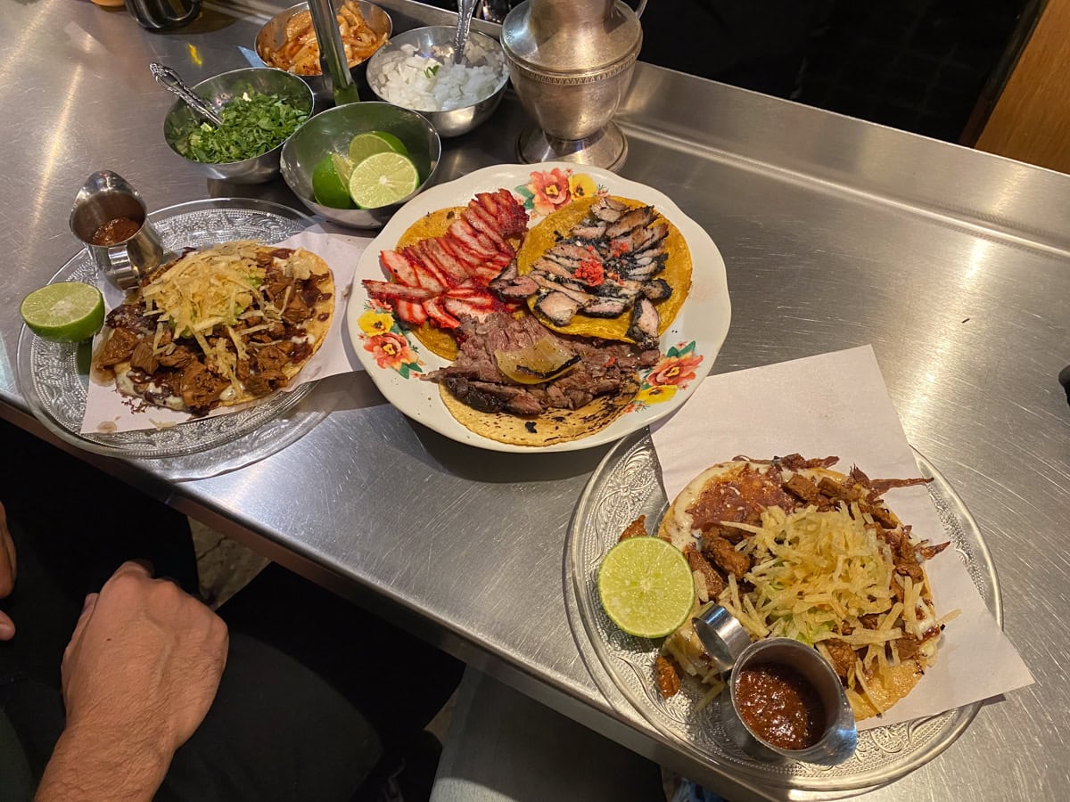 A close-up of three plates of tacos on a metal counter, each topped with meats, shredded potatoes, and served with lime wedges and small metal cups of salsa. Bowls of chopped cilantro, onions, and other garnishes are in the background.