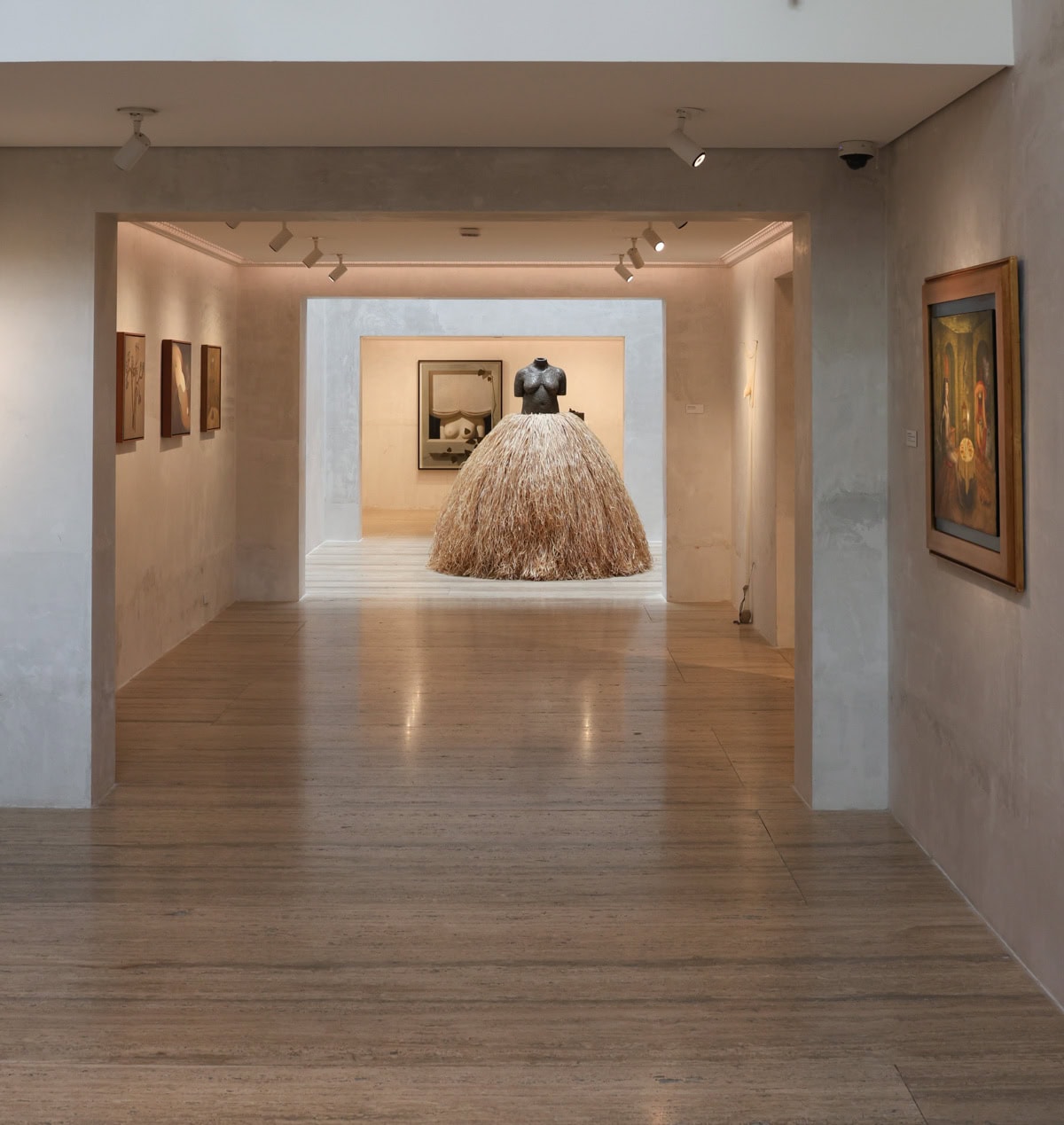 A contemporary gallery space featuring a striking sculpture of a female torso with a voluminous skirt made from straw-like material, displayed in a softly lit room framed by other artworks.